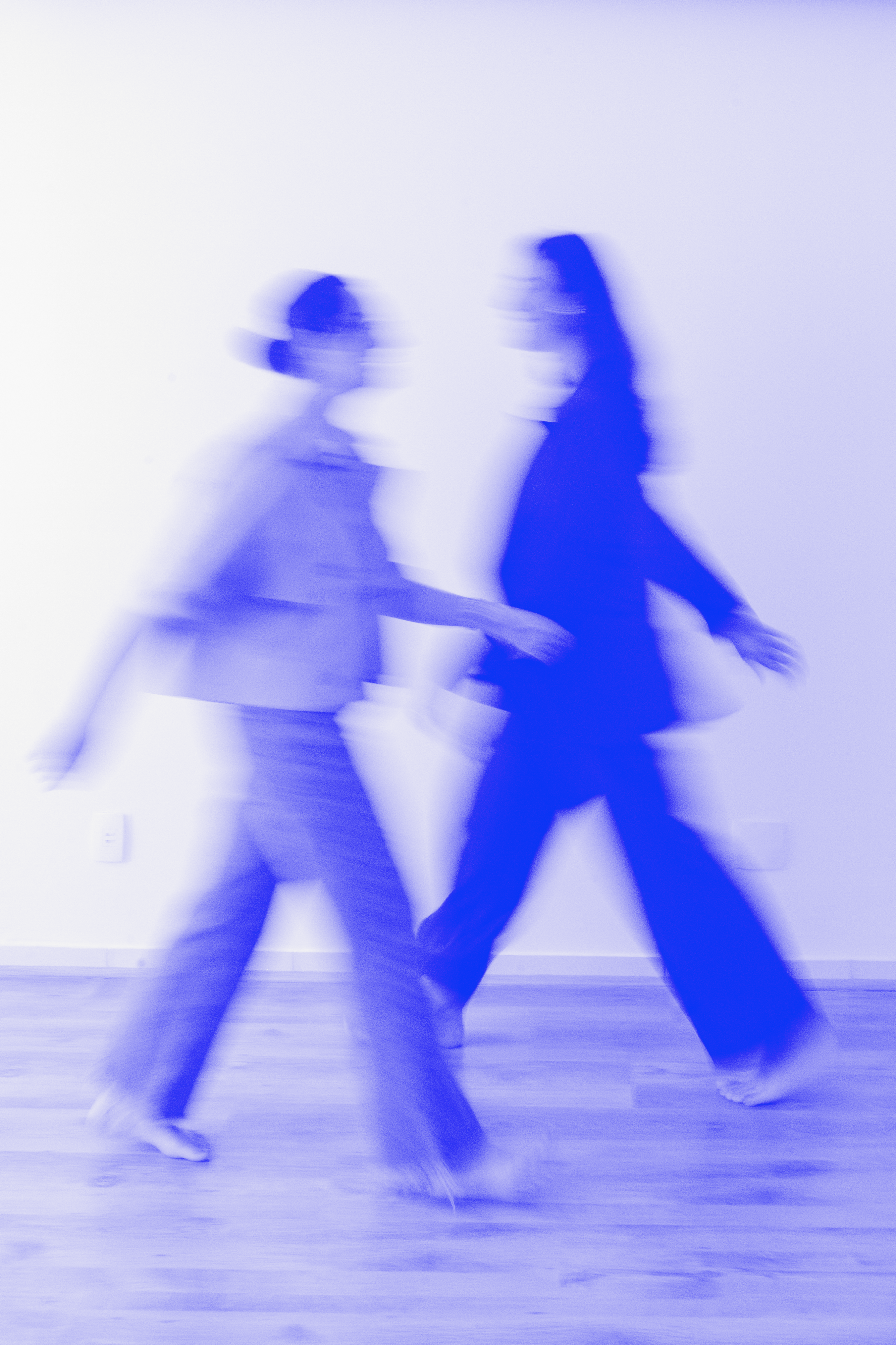 Duas pessoas em movimento, capturadas com efeito de desfoque, sobre um fundo branco com iluminação e tons predominantes de azul.
